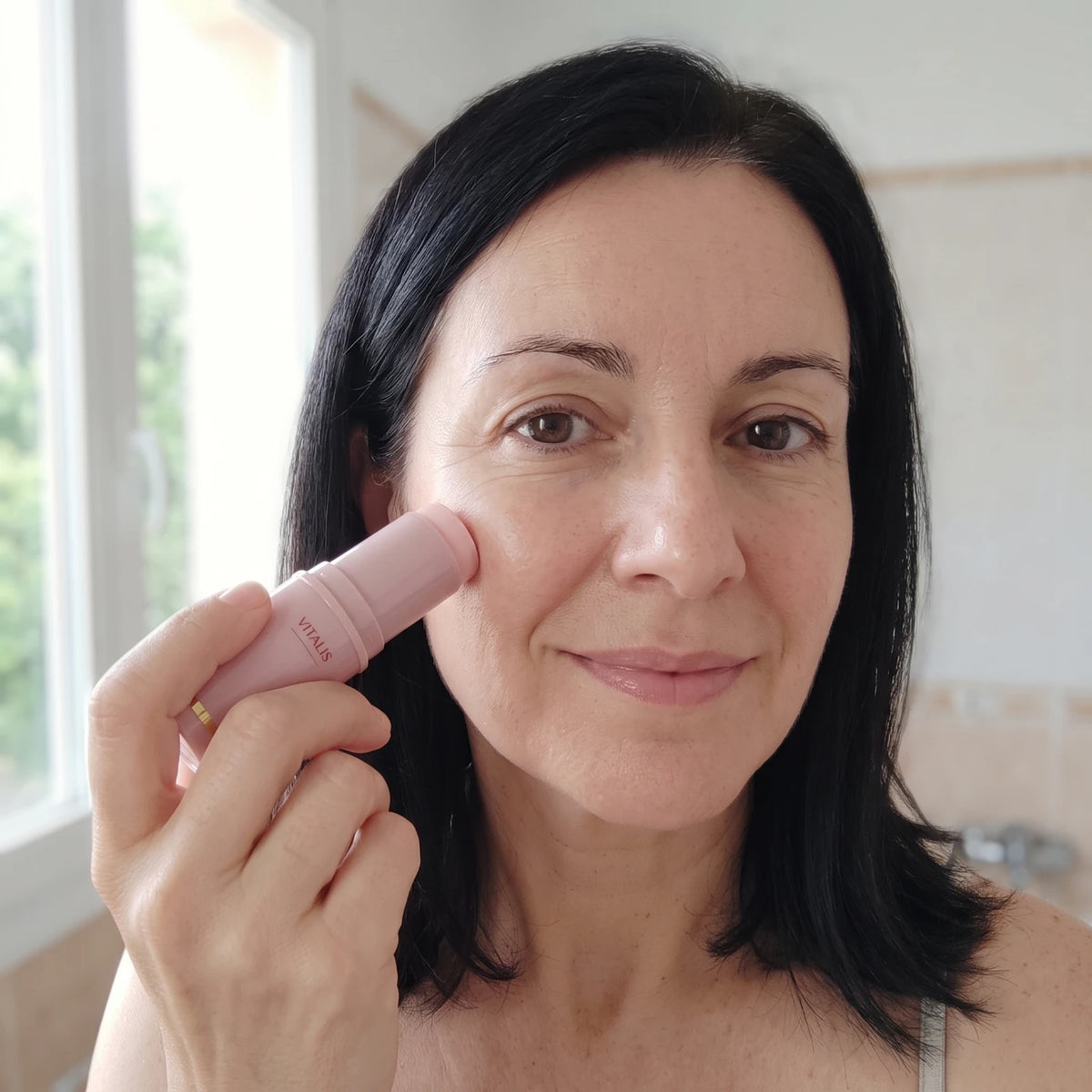 Mujer morena en casa, junto a la ventana, pasándose el stick por el pómulo